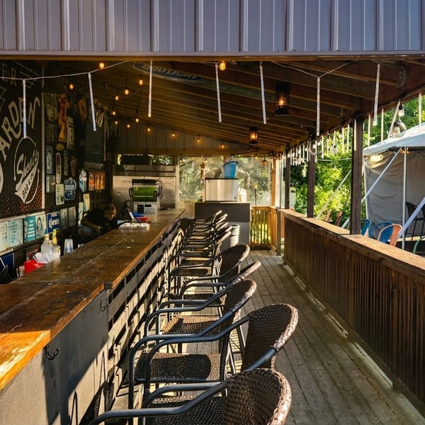 An outdoor bar with a long wooden counter, bar stools, string lights, and a tented seating area.