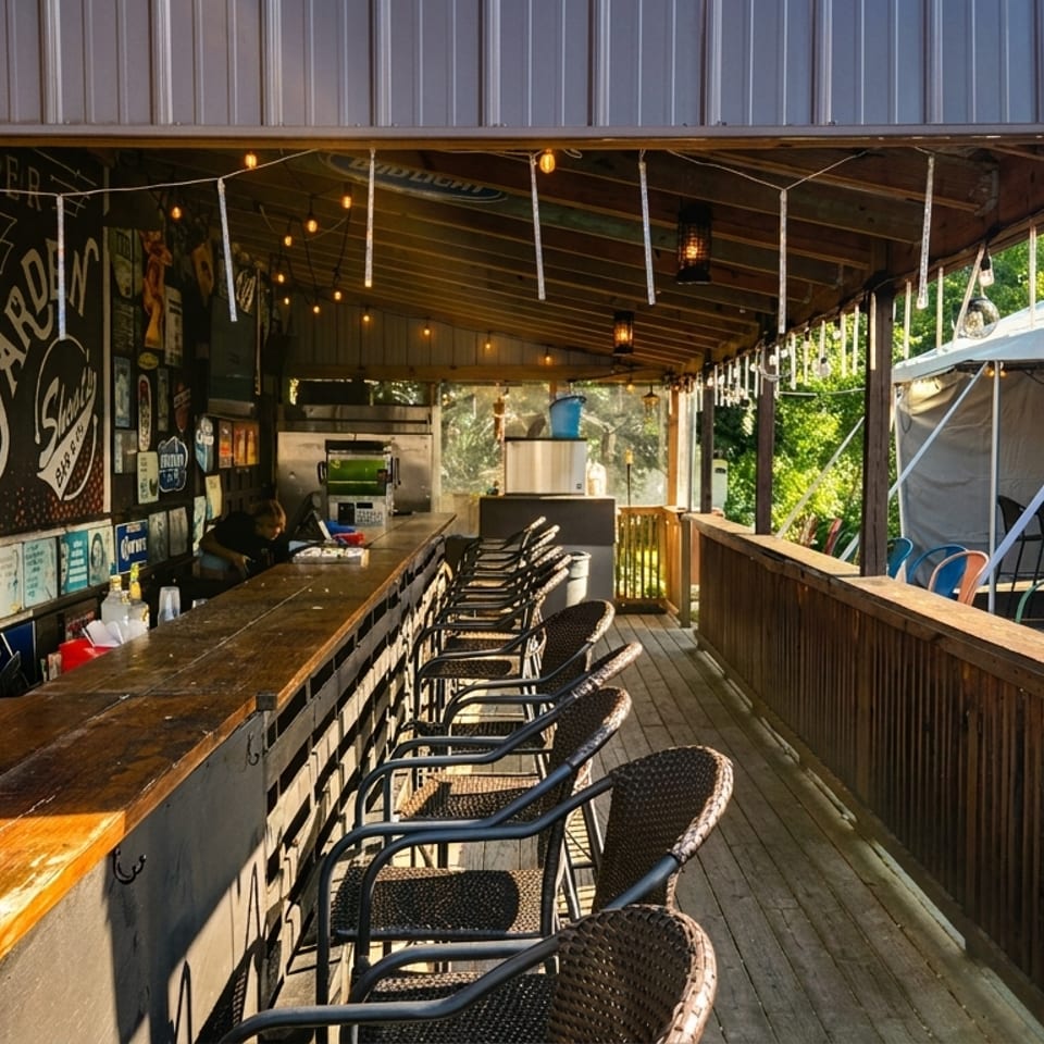 An outdoor bar with a long wooden counter, bar stools, string lights, and a tented seating area.