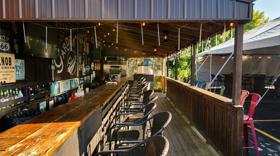 An outdoor bar with a long wooden counter, bar stools, string lights, and a tented seating area.