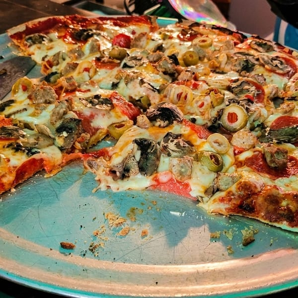 A partially eaten pizza with pepperoni, sausage, mushrooms, and olives on a metal pan.