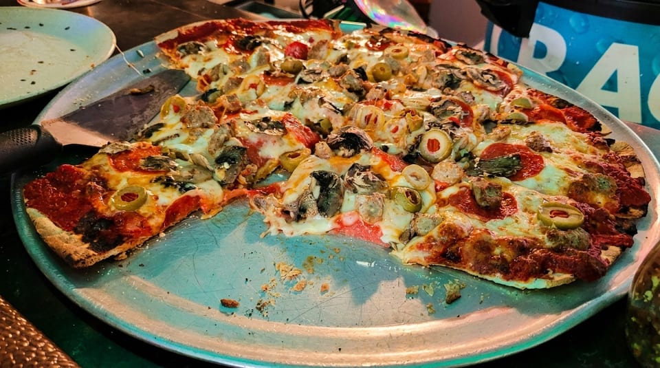 A partially eaten pizza with pepperoni, sausage, mushrooms, and olives on a metal pan.