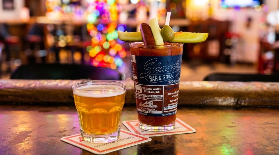 A Bloody Mary garnished with sausage, celery, and pickles next to a beer on a bar.