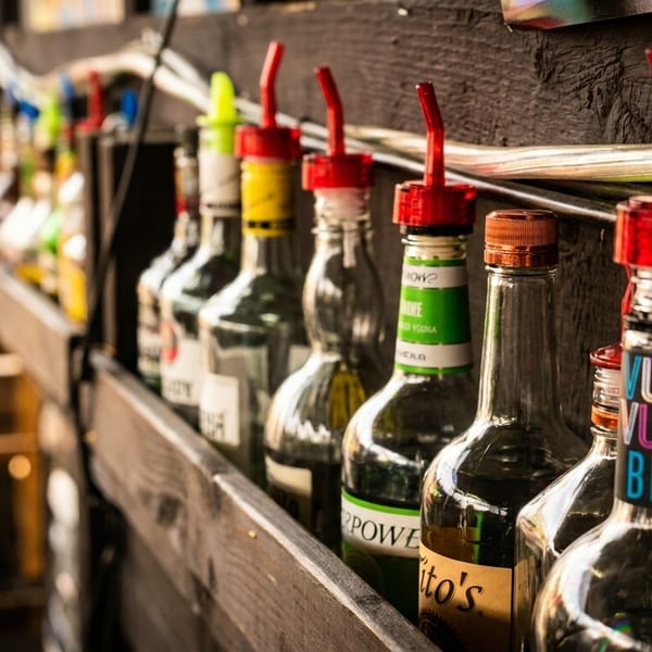 A row of liquor bottles with pour spouts on a wooden shelf at a bar.