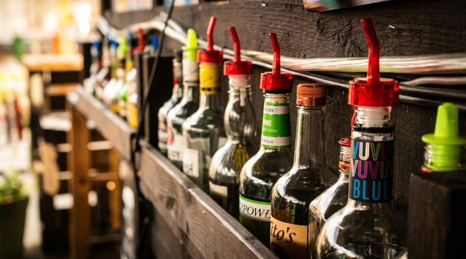 A row of liquor bottles with pour spouts on a wooden shelf at a bar.