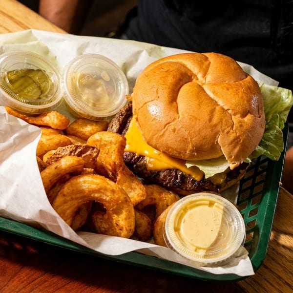 A cheeseburger with lettuce, curly fries, and dipping sauces in a green basket on a wooden table.