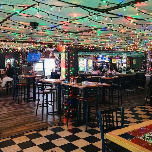 A lively bar interior with colorful string lights, patrons, high-top tables, and a checkered floor.
