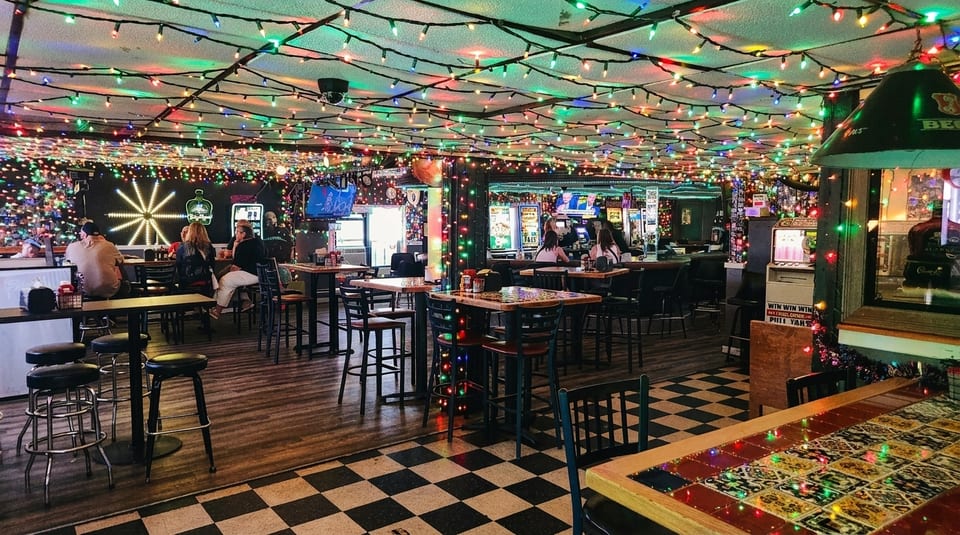A lively bar interior with colorful string lights, patrons, high-top tables, and a checkered floor.